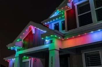 A house at night is decorated with multicolored LED lights along the roofline, displaying red, green, and blue hues. The lights illuminate the exterior and give a festive appearance.