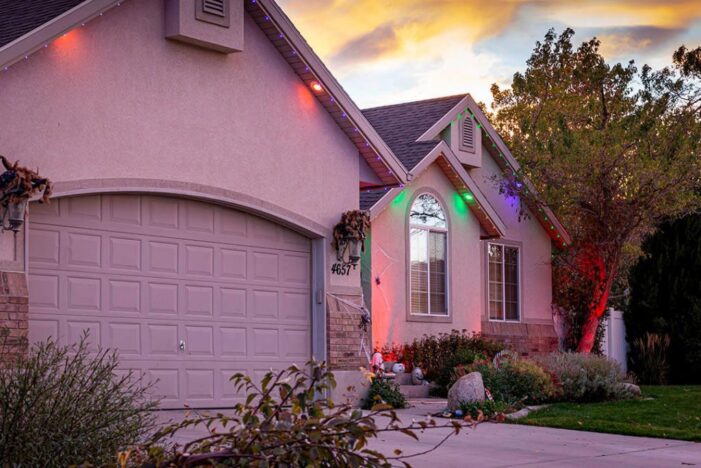 A suburban house at sunset with a two-car garage. The house is decorated with colorful holiday lights along the roof and front window. Trees and bushes surround the house, and the driveway is empty.