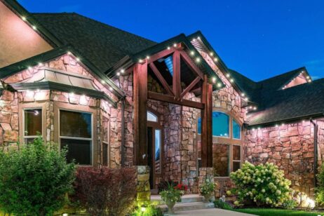 A large stone house with wooden beams and outdoor lighting, illuminated at dusk. The entrance features steps, plants, and large windows; the roof is dark and the sky is clear and blue.