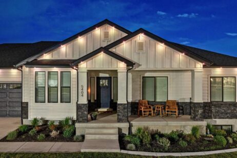 A modern, single-story house at night with exterior lights on. The house has white siding, dark trim, a small porch with two wooden chairs and a table, landscaped front yard, and the number 5404 by the door.
