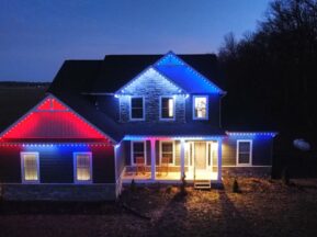A two-story house at dusk is decorated with red, white, and blue lights along the roofline. The house is illuminated, and trees and open land are visible in the background.