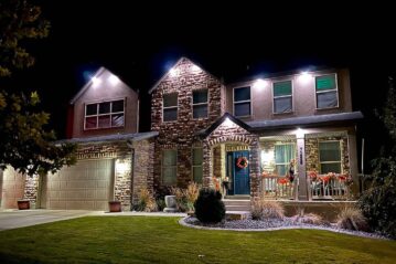 A two-story house with stone and stucco exterior is lit up at night. It has a front porch decorated with orange fall garlands and a wreath on the blue door. The lawn is neatly trimmed, with some shrubs and potted plants.