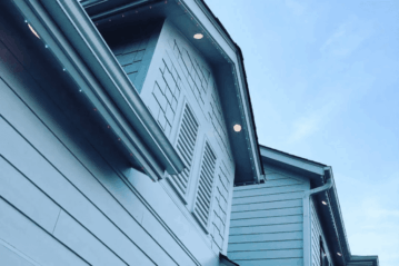 Close-up view of the upper exterior of a light blue house with horizontal siding, decorative shutters, and recessed lights under the eaves against a clear blue sky.
