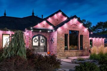 A brick house at dusk is decorated with pink and white lights along the roofline, featuring arched double doors, large windows, wreaths, and some landscaping with bushes and ornamental grass in the foreground.