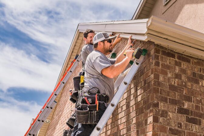 Two workers on ladders install or repair a gutter on the roof edge of a brick house. They wear grey shirts and tool belts, working under a partly cloudy sky.