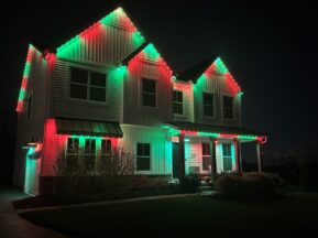 A two-story house at night is decorated with red and green lights along the roofline, creating a festive holiday appearance. Shrubs and a driveway are visible in the front yard.
