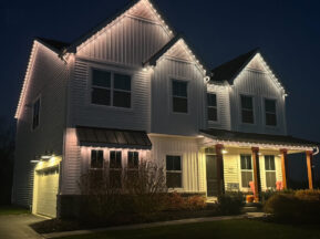 A two-story white house at night is illuminated by white string lights along the roofline. The porch has wooden columns, two orange chairs, a pumpkin, and bushes in front. The garage is to the left.