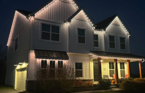 A two-story white house at night is illuminated by white string lights along the roofline. The porch has wooden columns, two orange chairs, a pumpkin, and bushes in front. The garage is to the left.