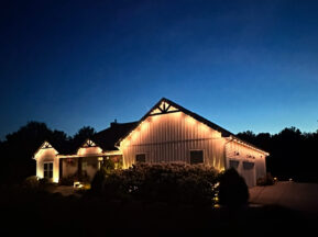 A house is decorated with glowing string lights along the roofline at dusk, surrounded by bushes and trees, with a deep blue sky in the background.