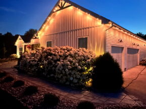 A white house with a garage is illuminated by warm string lights along the roof at dusk, surrounded by blooming bushes and landscaping stones under a deep blue sky.