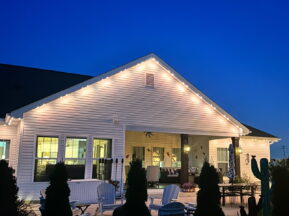 A cozy backyard patio at dusk features white outdoor chairs, a table, potted plants, and string lights illuminating the roofline of a white house against a deep blue evening sky.