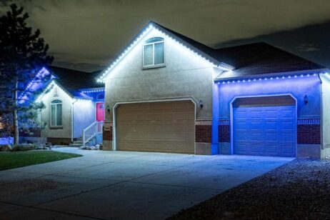 A single-story house with a double garage, lit by blue and white LED lights along the roofline at night. The driveway and part of the lawn are visible in the foreground.