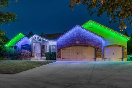 A single-story brick house at night with green and white LED lights outlining the roof, three garage doors, and a well-kept front yard with trees and bushes visible.