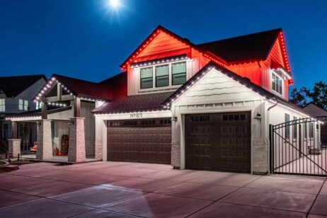 A modern two-story house at night is decorated with red and white lights along the roofline. The house has three garage doors, gabled roofs, a gated driveway, and a bright full moon overhead.