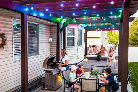 A woman grills food on a patio while children eat at a nearby table. In the background, several people and a dog relax in a raised hot tub. Colorful string lights hang above the deck, creating a festive atmosphere.