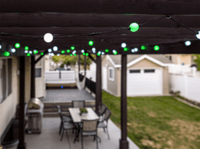String lights with green and white bulbs hang from a pergola over a patio with a dining table and chairs. In the background, there is a lawn, a barbecue grill, and a detached garage.
