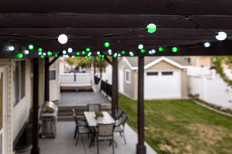 String lights with green and white bulbs hang from a pergola over a patio with a dining table and chairs. In the background, there is a lawn, a barbecue grill, and a detached garage.