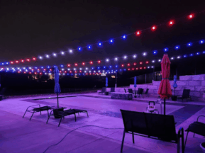 A swimming pool at night is illuminated by strings of red, white, and blue lights overhead. Lounge chairs and closed umbrellas line the poolside, creating a festive yet quiet atmosphere.