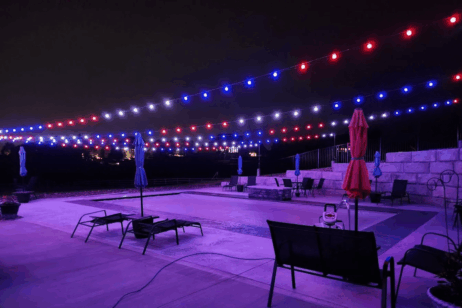A swimming pool at night is illuminated by strings of red, white, and blue lights overhead. Lounge chairs and closed umbrellas line the poolside, creating a festive yet quiet atmosphere.