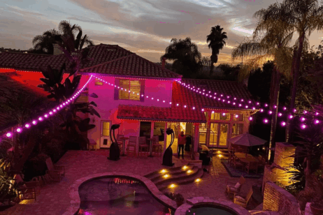 A backyard patio at dusk features a pool, palm trees, and a two-story house decorated with purple string lights, creating a festive ambiance. Outdoor seating and a dining area are visible.