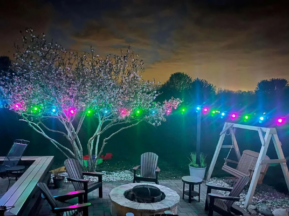 A backyard patio at night features a fire pit surrounded by chairs, a wooden swing, blooming trees, and multicolored string lights, with a cloudy sky illuminated by city lights in the background.
