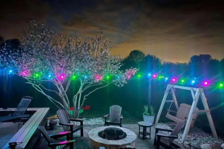 A backyard patio at night features a fire pit surrounded by chairs, a wooden swing, blooming trees, and multicolored string lights, with a cloudy sky illuminated by city lights in the background.