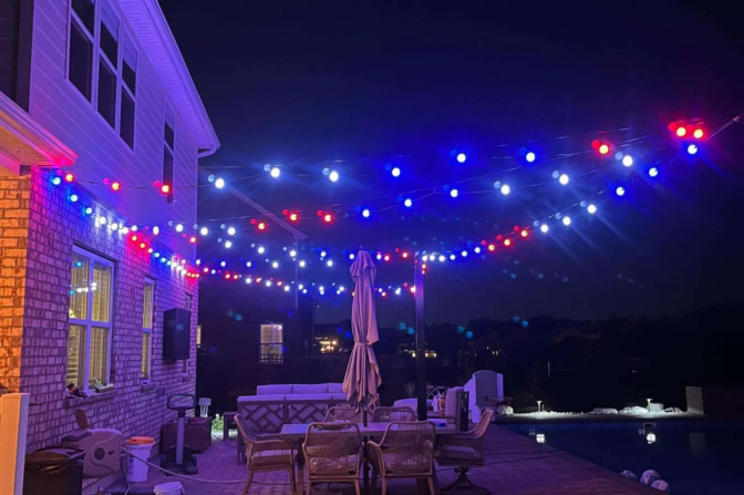 A backyard patio at night is decorated with strings of red, white, and blue lights. Patio furniture, including chairs and a closed umbrella, sits near a pool. The house and pool are softly illuminated.
