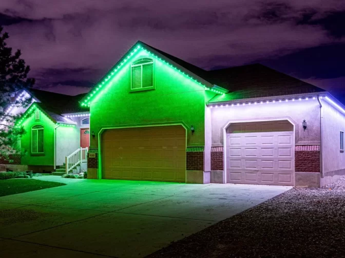 A suburban house at night features green and white LED lights along the roofline, illuminating the garage doors and front entrance. The driveway and sky are visible, with some clouds overhead.