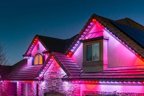 A house at dusk decorated with pink, purple, and white LED lights outlining the roof and gables, creating a festive and colorful appearance against the evening sky.