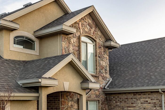 Close-up of a suburban house exterior featuring stone and beige stucco siding, gray shingled roof, and multiple gabled sections with white-trimmed windows. Sky is clear in the background.