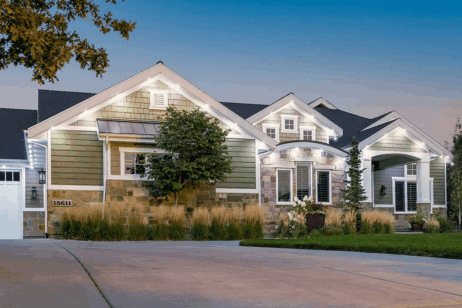 A modern suburban house at dusk with green siding, stone accents, white trim, large front windows, a manicured lawn, shrubs, and exterior lights illuminating the entrance and facade.