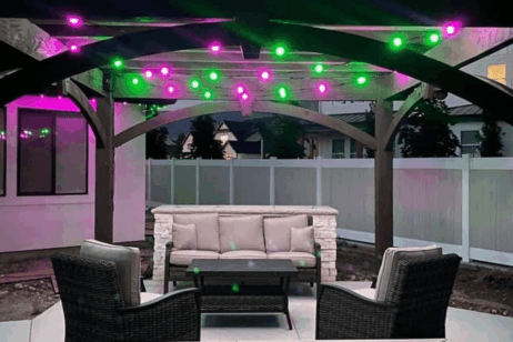 A covered outdoor patio with a cushioned sofa, two wicker chairs, and a coffee table, illuminated by pink and green string lights hanging from the pergola overhead.