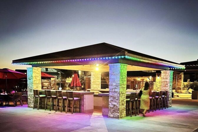 Outdoor bar with stone pillars and a dark roof, outlined by colorful string lights at dusk. Barstools surround the bar, and a person is walking nearby. Patio umbrellas and seating are visible in the background.