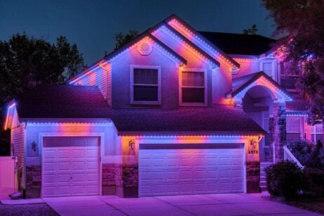A two-story house at night is decorated with bright purple, pink, and blue LED lights outlining the roof, garage, and entryway, creating a colorful and festive appearance.