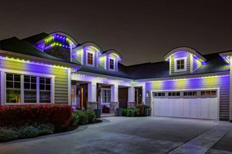A two-story suburban house at night with white and purple LED lights outlining the roof and dormer windows, creating a festive look. The driveway and front yard are visible in the foreground.