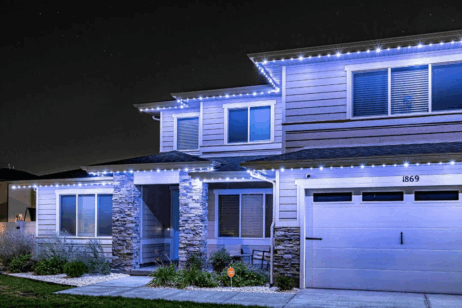 A modern two-story house at night, outlined with bright white LED lights along the roofline. The house has a two-car garage, stone pillars, and well-kept landscaping. The address number 1869 is visible above the garage.