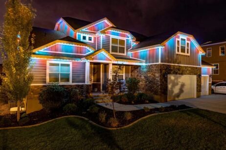 A two-story house at night is decorated with colorful LED strip lights along the roofline, illuminating the exterior with blue, red, and white hues. The house has stone accents, a porch, and a well-kept front yard.