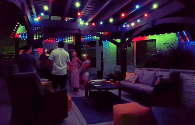 A group of people chat and relax under a pergola decorated with colorful string lights at night. Outdoor couches and a table with cups create a cozy, festive atmosphere.