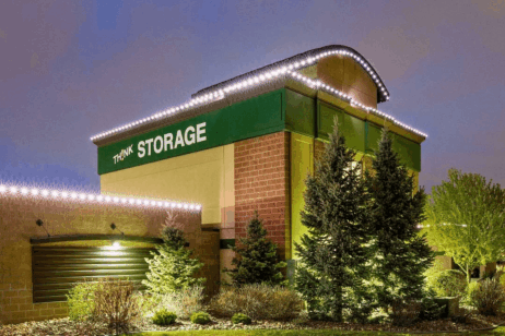 A well-lit storage facility at dusk with a green sign reading “THINK STORAGE,” surrounded by trees, shrubs, and exterior string lights along the roofline.